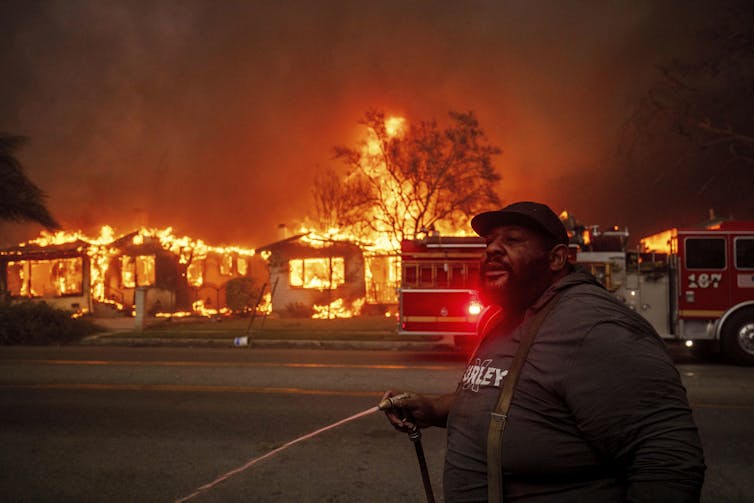 A man holds a fire hose to try to safe a property as a row of homes behind him burn