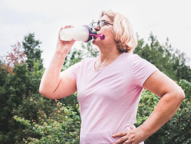 An older woman wearing a pink shirt and standing outdoors drinks out of a shaker bottle used for protein or creatine shakes.