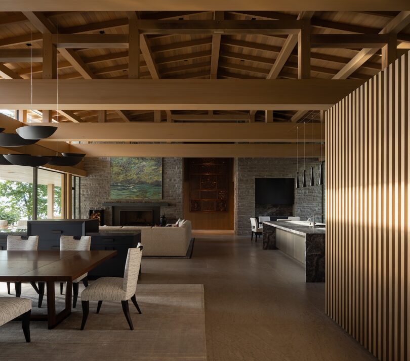 Modern open-concept living and dining area with exposed wooden beams, stone walls, contemporary furniture, and large windows letting in natural light.