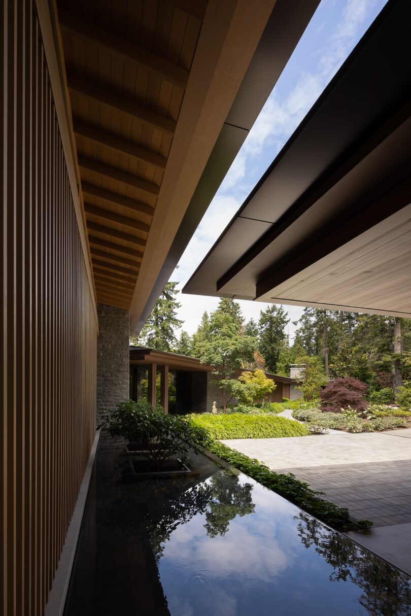 Modern house exterior with wooden and stone elements, overhanging roofs, a reflective water feature, and landscaped garden under a partly cloudy sky.