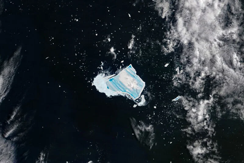 Aerial view of a large iceberg with bright blue stripes floating in dark ocean water, surrounded by smaller ice fragments and patches of white clouds.