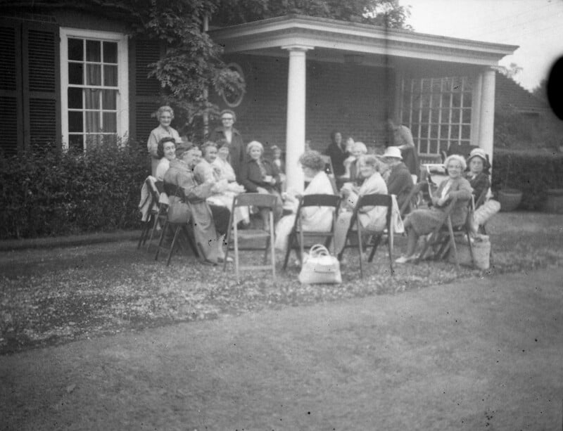 A group of women sit on folding chairs around tables outdoors, in front of a house with large windows and a covered porch, appearing to enjoy a social gathering. Some hold cups and bags are placed nearby.