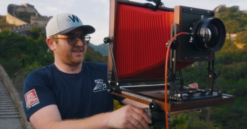 A man wearing glasses and a hat sets up a large-format camera with a red bellows, outdoors near the Great Wall of China, with lush green hills in the background.