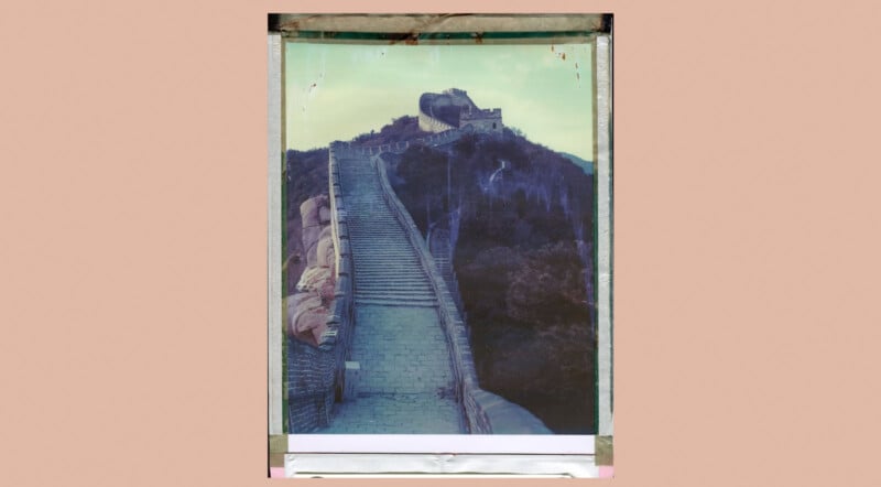 A faded photograph of the Great Wall of China winding up a hilly landscape, bordered by tape on all sides, set against a plain light brown background.