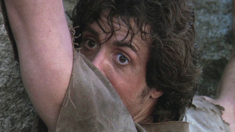 John Rambo grabbing onto a mountainside and looking over his shoulder in First Blood