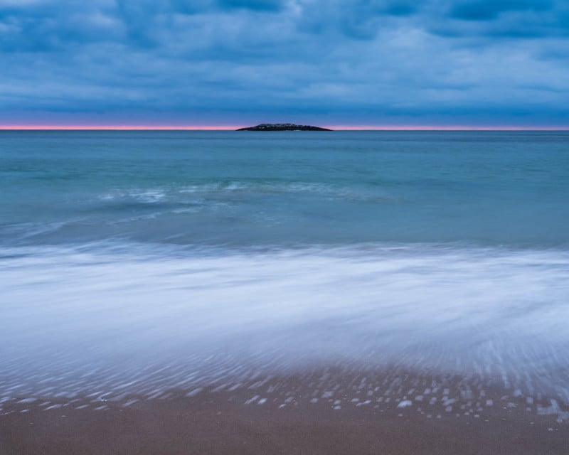 Waves gently wash onto a sandy beach under a cloudy blue sky, with a small island visible on the horizon and a thin line of pink light at sunset or sunrise.