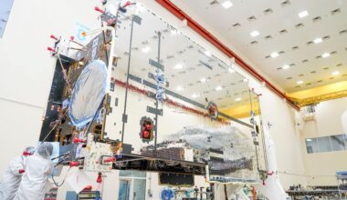 The primary structure of Gateway’s Power and Propulsion Element stands inside a high-bay cleanroom at Lanteris Space Systems in Palo Alto, California. The large rectangular structure is covered in reflective silver-colored panels. Two technicians in white cleanroom suits work near the base of the structure.
