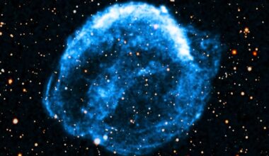A glowing blue nebula appears as a circular, cloud-like formation against a dark background filled with scattered stars. The nebula glows intensely, with wispy edges and bright, varying light within it.