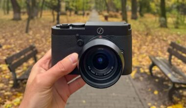 A hand holds a black Leica camera in the center of a park pathway lined with benches and autumn trees, with yellow leaves scattered on the ground.