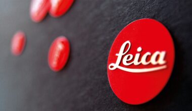 A close-up photo of multiple red circular badges on a black surface, with the frontmost badge displaying the white cursive "Leica" logo. The background badges are out of focus.