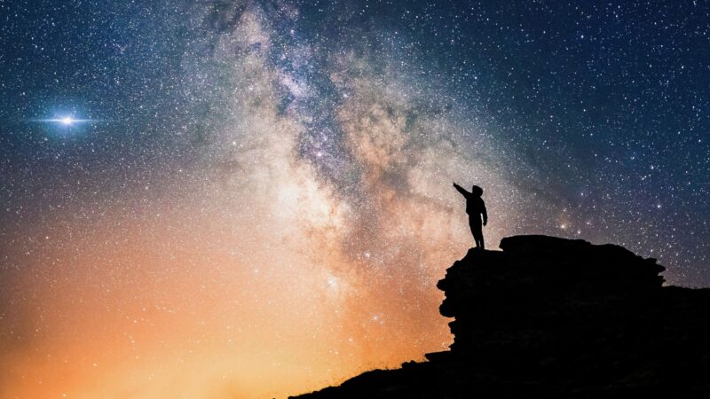 Lone person standing on top of rock, pointing towards the vivid cloudy Milky Way and the vast expanse of universe.