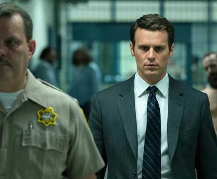 An agent in a suit and tie is escorted by a guard through a prison as inmates look on in Mindhunter.
