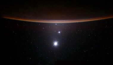 Earth's crescent looks like an orangey sliver with a backdrop of space below it. The moon shines as a bright ball of light while Jupiter and Venus are arranged as bright dots above it.