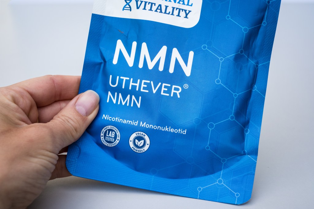 A person holds a blue package of Uthever NMN Nicotinamide Mononucleotide powder, labeled "Lab Tested for Purity & Potency" and "Vegan Friendly."
