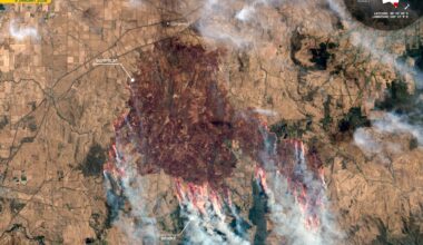 A brown landscape is scarred dark red as streaks of orange flames are seen around the edges of the dark area with streams of smoke moving toward the bottom of the image