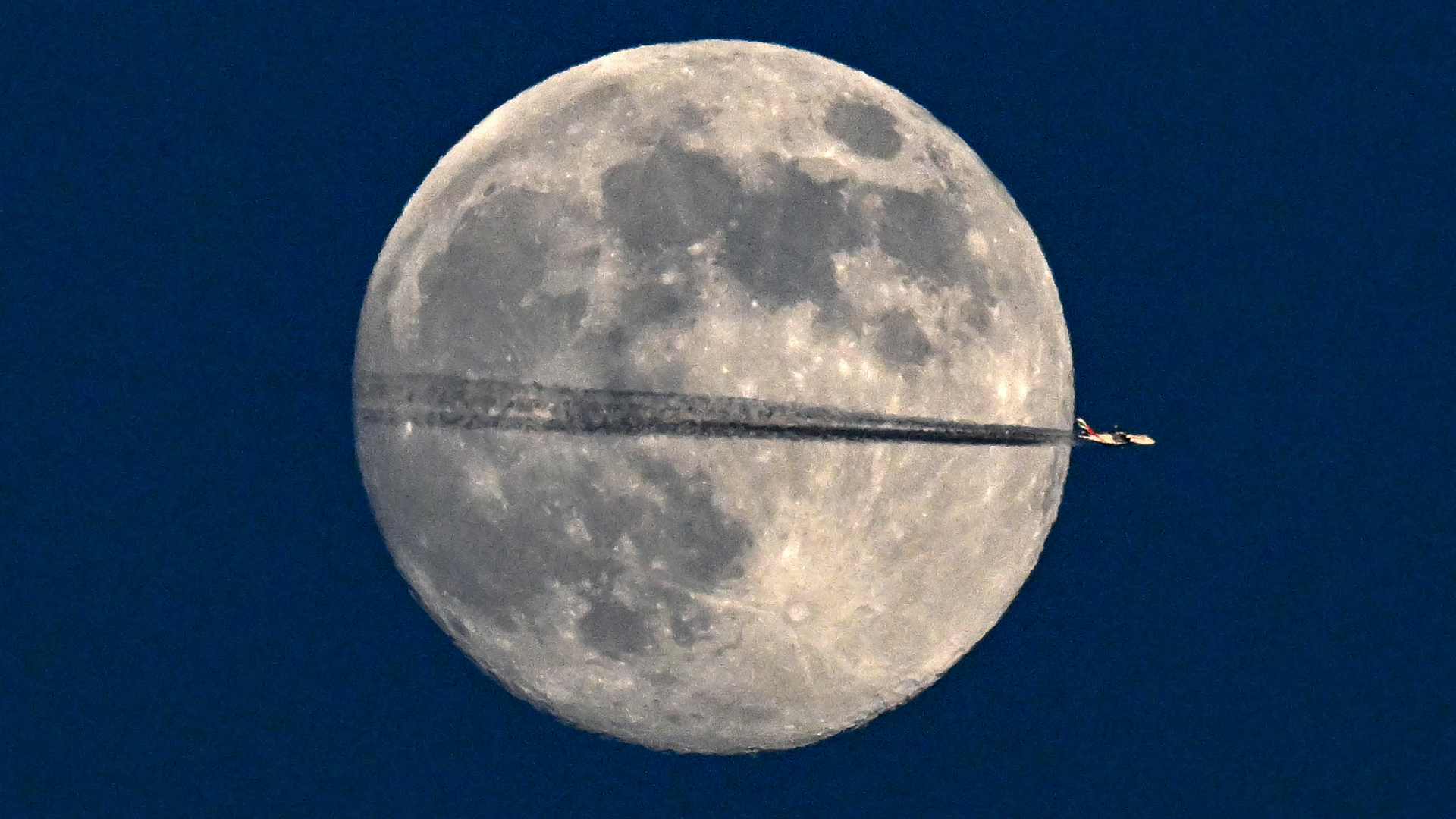 A commercial jet is pictured in profile having crossed the lunar disk in the night sky. The jet has left a dark exhaust trail across the moon's equator, where the dark-grey expanses of lava plains and craters mark its surface.