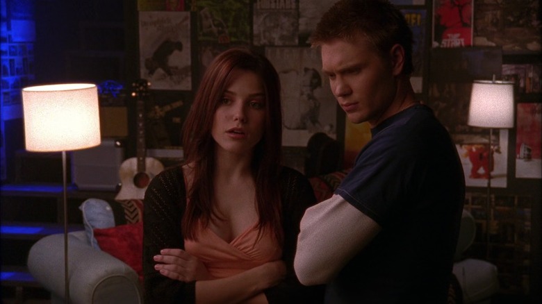 Sophia Bush as Brooke with arms crossed and music posters behind her standing next to Chad Michael Murray with arms crossed and furrowed brow in One Tree Hill