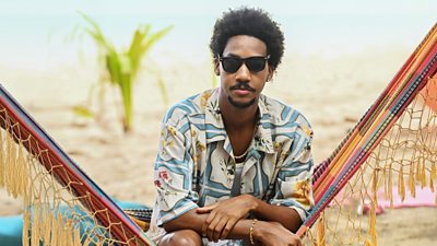Shaquille Ali-Yebuah in character as Officer Sebastian Rose. Wearing sunglasses and a colourful shirt, he sits on a hammock on a beach. 