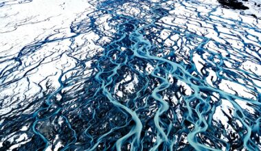 Aerial view of a glacial river meandering through a snowy landscape, surrounded by towering mountains and blue sky
