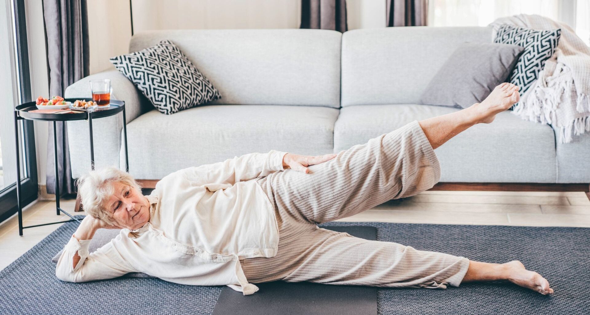 woman dressed in cream loose top and trousers lying on her side facing the camera with her top leg raised. she's on a grey carpet with a light grey sofa behind her.