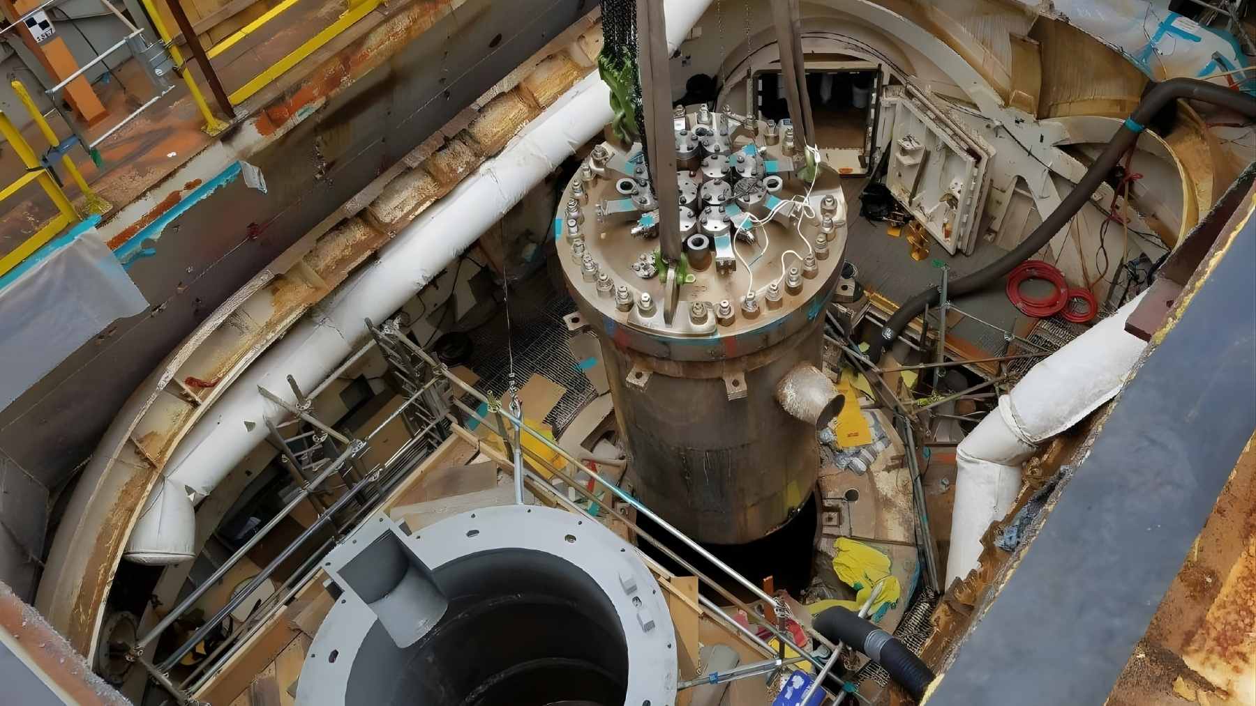 A large cylindrical naval nuclear reactor component is being lifted inside a shipyard work area during maintenance or removal.