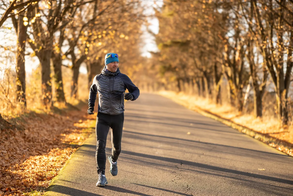 Smiling middle-aged man dressed sporty in black leggings and blue jacket running through forest full of fallen golden leaves on beautiful autumn day.