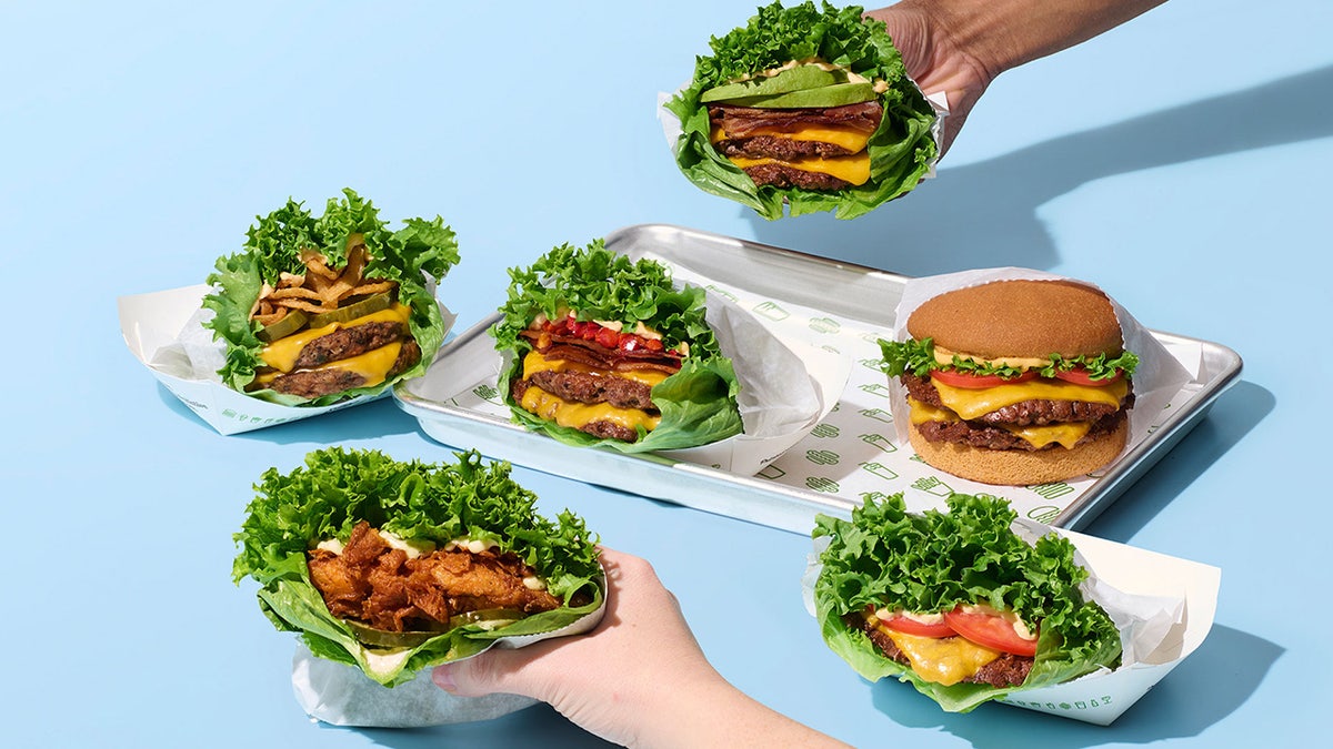 New lettuce-wrapped burger and chicken sandwich items part of Shake Shack's newly released "Good Fit Menu." Two hands holding lettuce-wrapped sandwiches with others including double cheeseburgers on trays and in wrappers alongside.