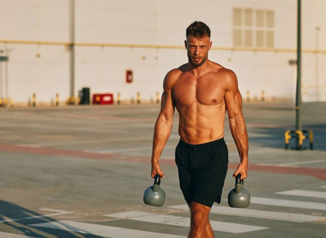 Muscular athlete walking and carrying two kettlebells in a parking lot
