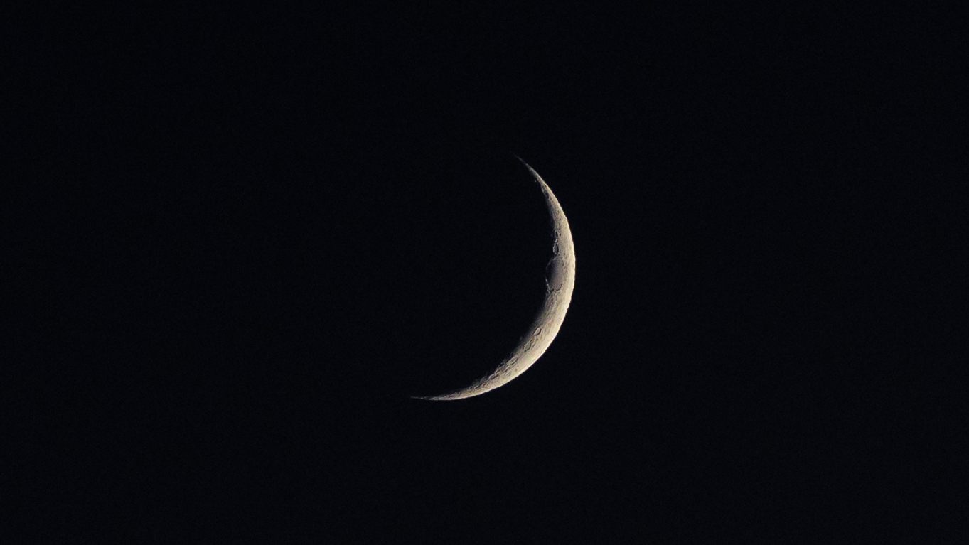 A photograph of a thin waxing crescent moon against a black night sky. A large oval impact crater dominates the upper limb of the sunlit surface.