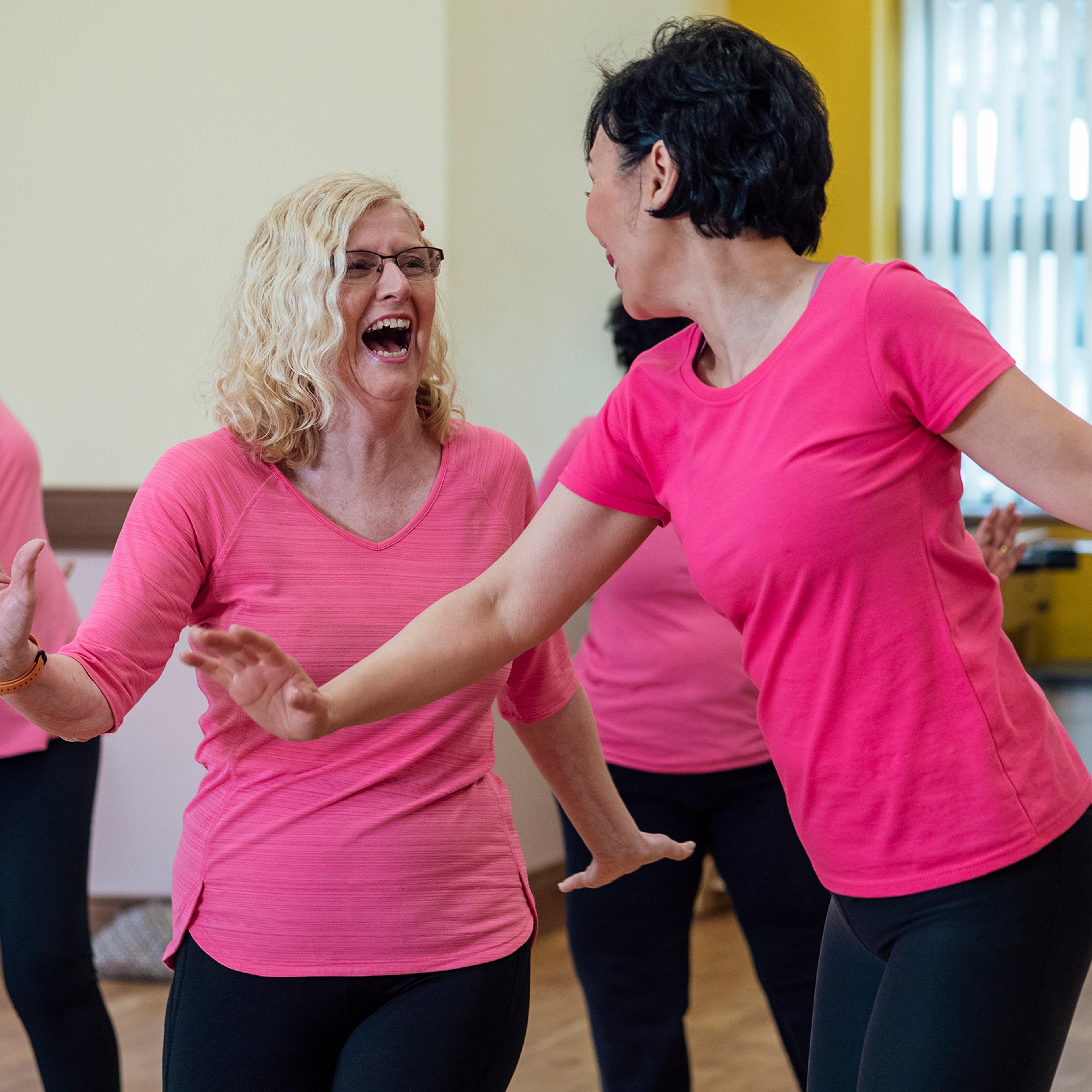 Women laughing and dancing in a studio, all wearing pink tops and black leggings.