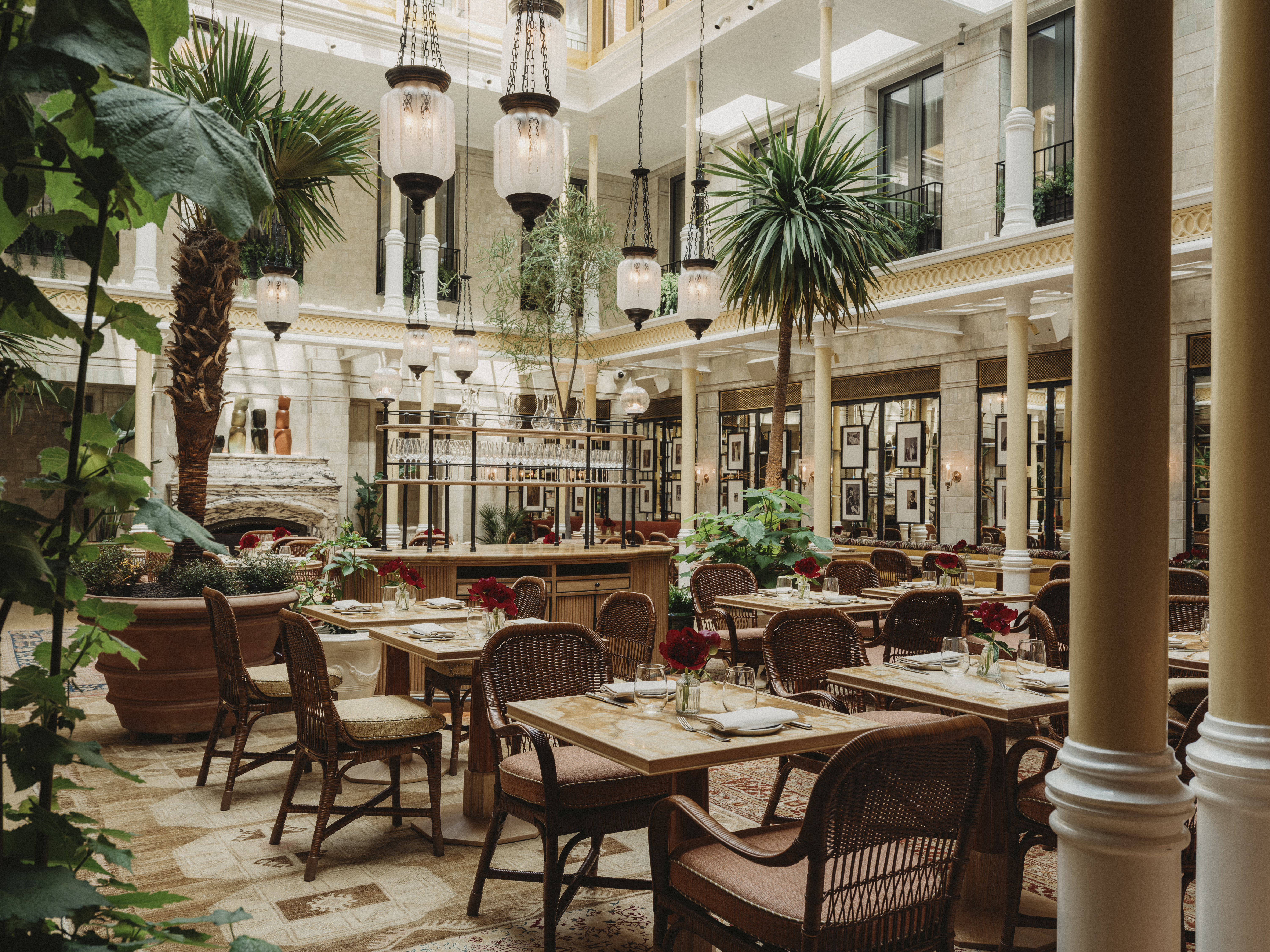 A naturally lit indoor hotel atrium filled with palm trees, wood, fabric, and marble furnishings, a glamorous central bar with an Art Deco style, pendant glass and wrought iron chandeliers, and plenty of framed photographs hanging on its walls.