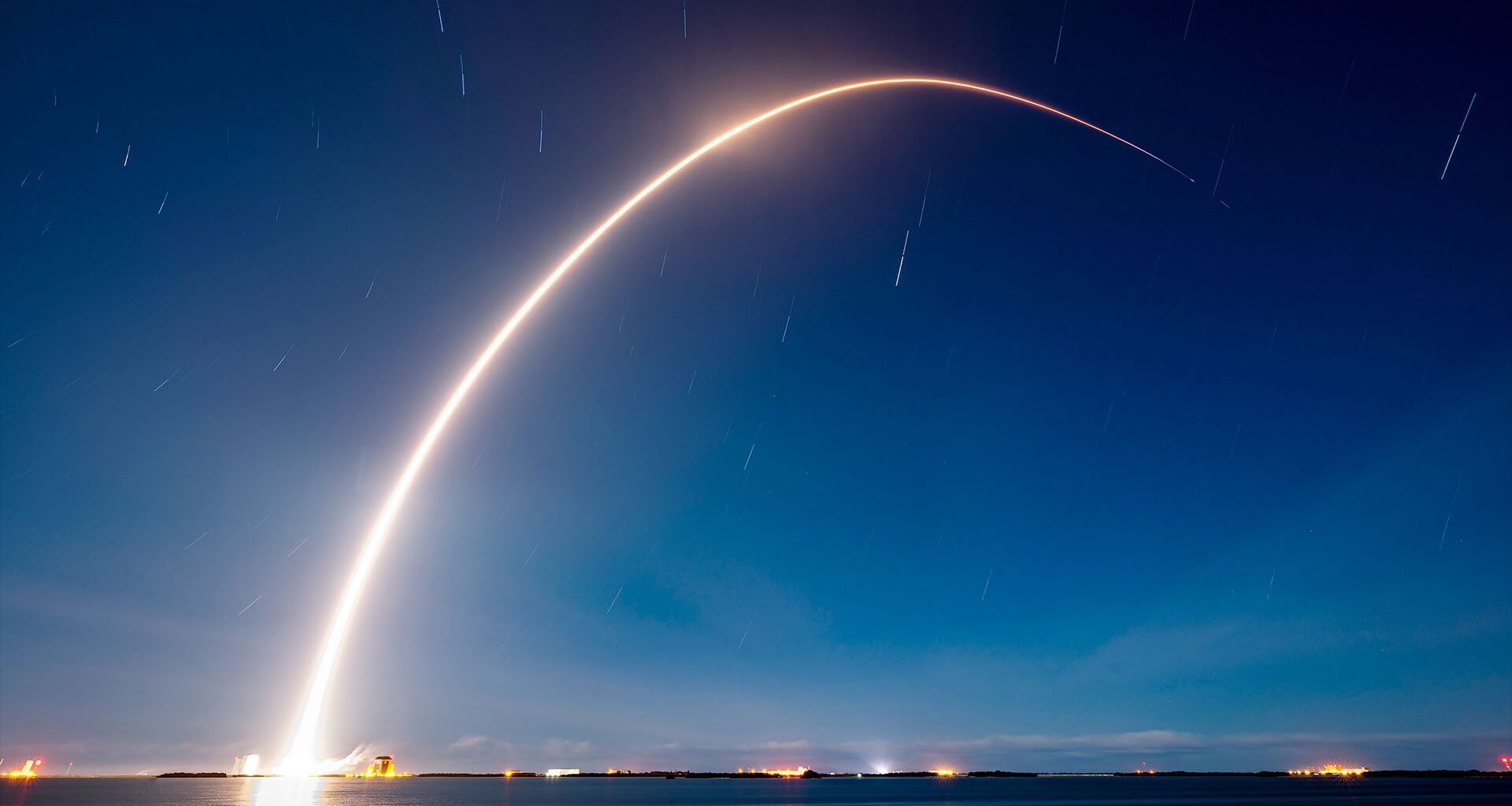A time lapse photo captures the break streak of a rocket launching into the early morning dark blue sky over a body of water