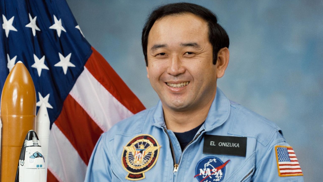 a man in a blue flight suit poses for a portrait beside an american flag and a model of a rocket