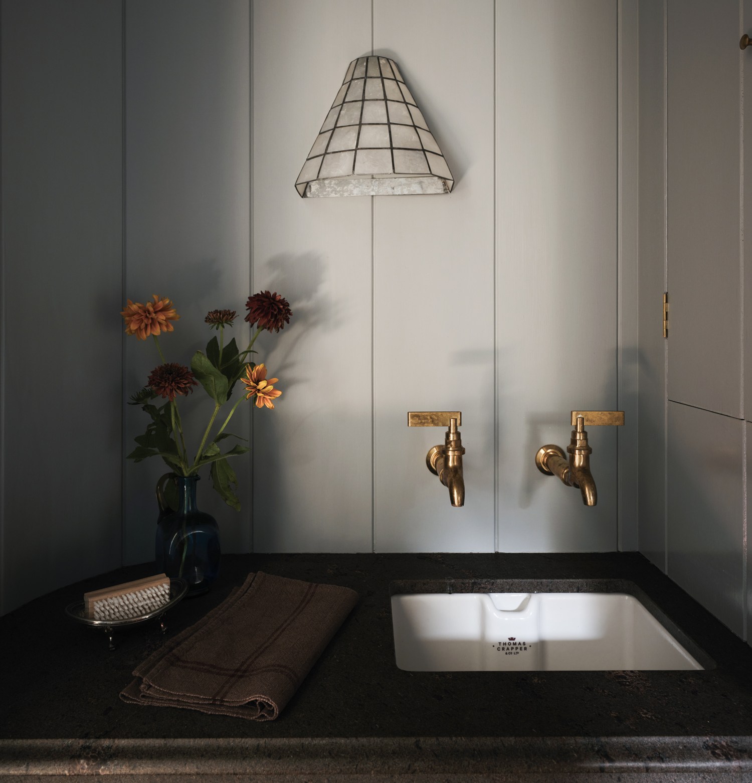 A small white sink set into a dark countertop, with two brass wall-mounted taps and a vase of flowers beside it.
