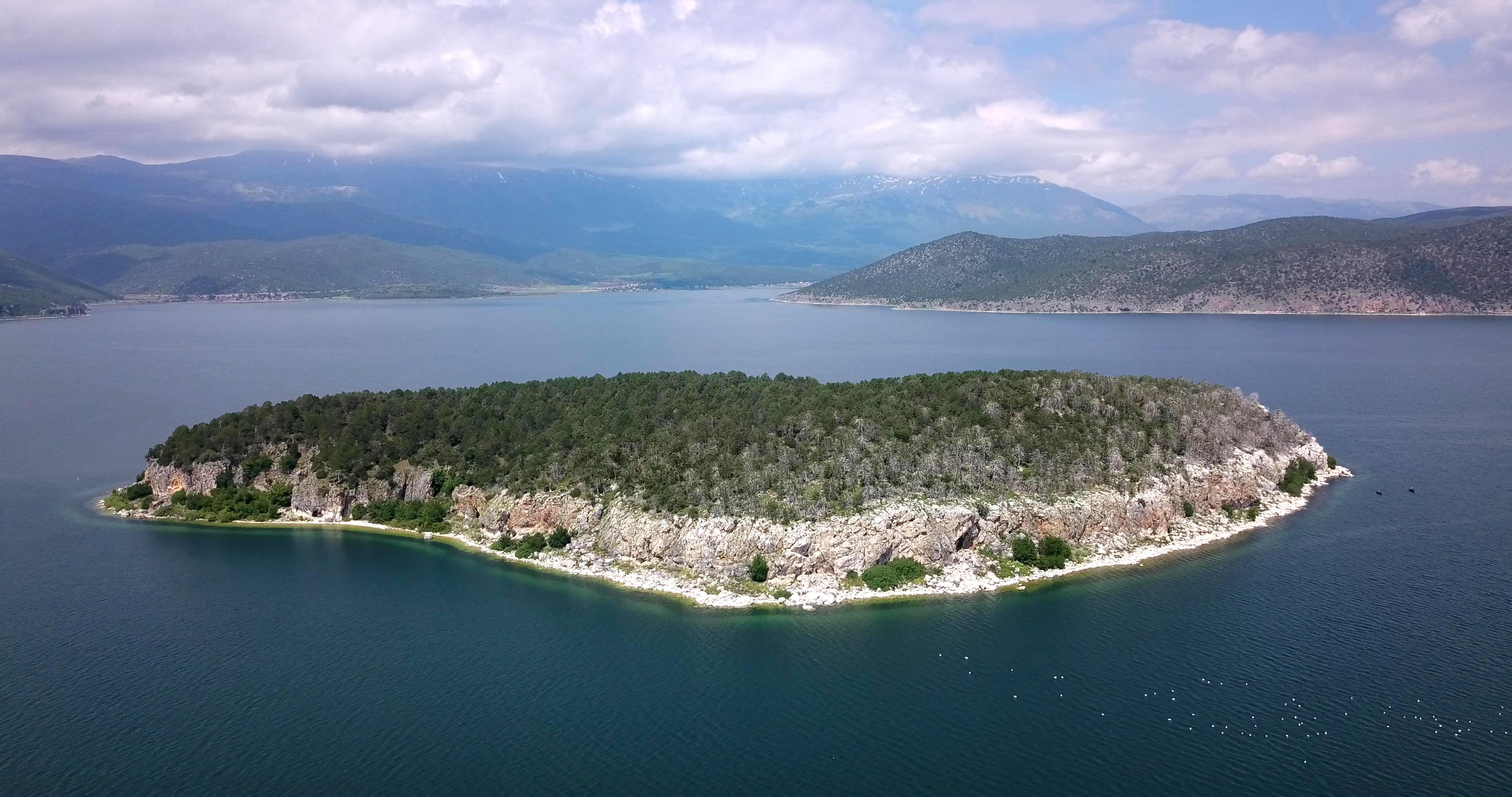 Aerial view of Golem Grad island in Lake Prespa, North Macedonia, with rocky shores and green trees, surrounded by blue water and distant mountains under a cloudy sky.