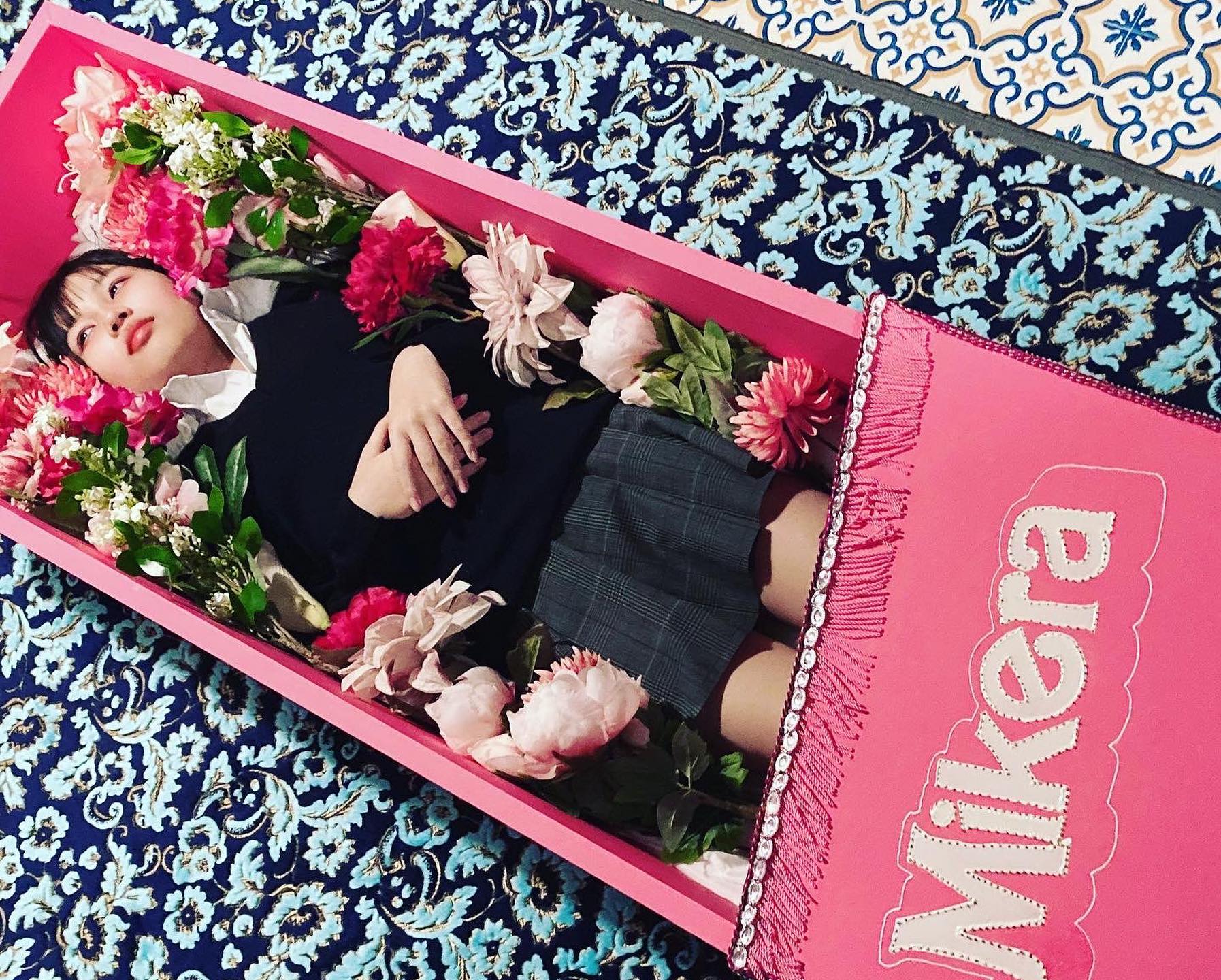 A young woman dressed in a school uniform, lying in a pink coffin filled with flowers, against a blue patterned rug.