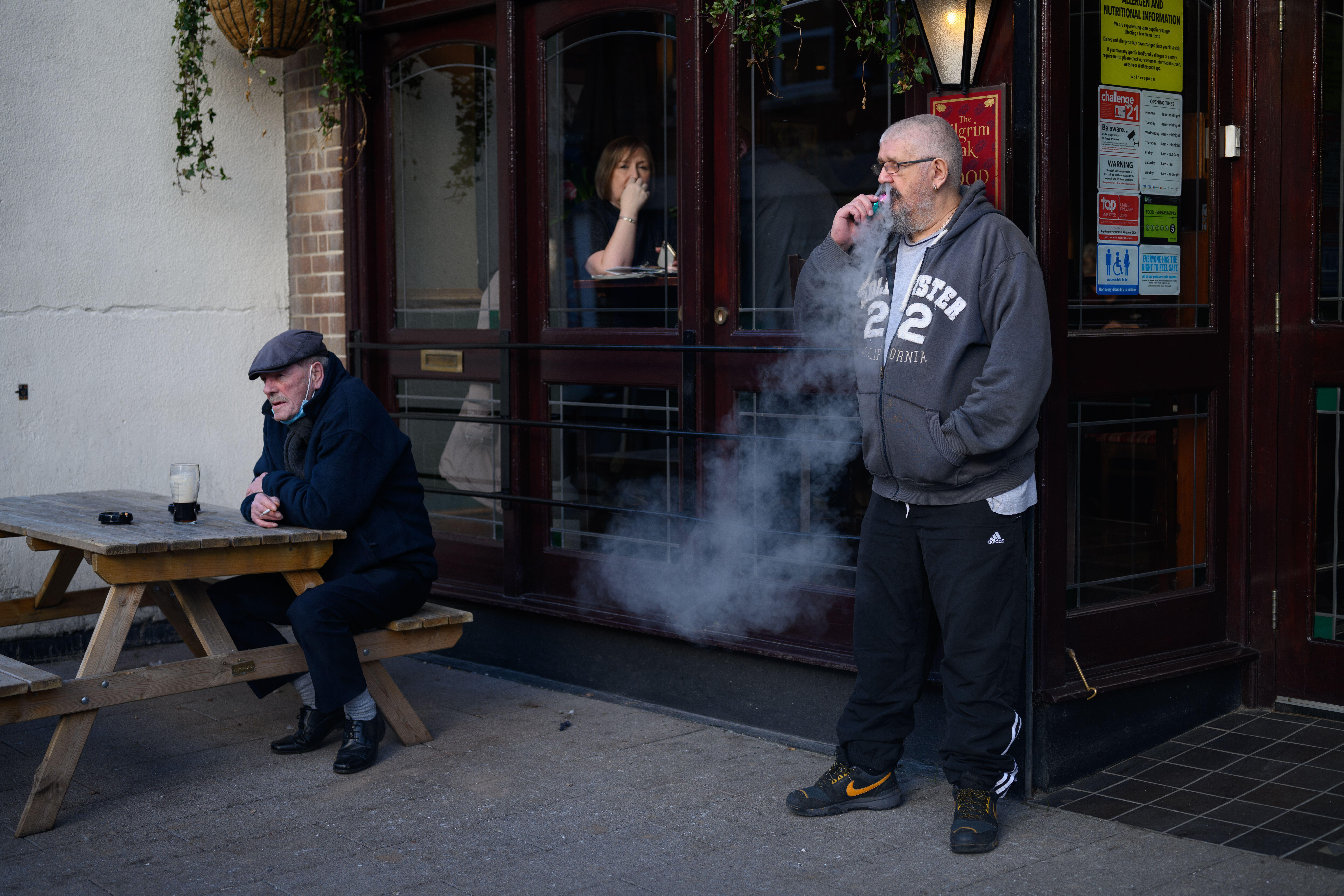 A man smoking outside a public house with a beer on a table next to another man, while a woman looks out from inside the pub in Hucknall, Nottinghamshire.