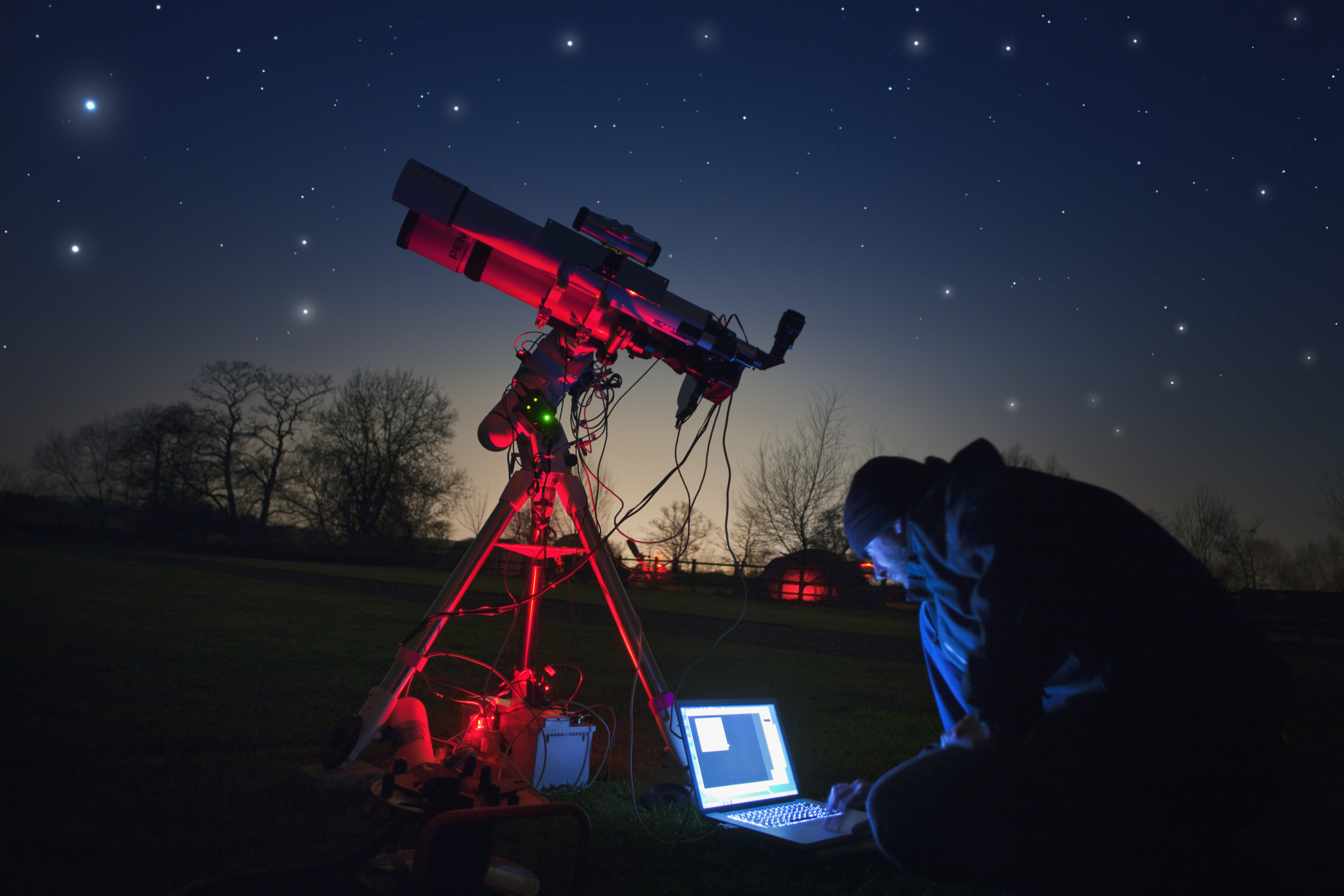 An amateur astronomer guides his red-lit telescope via a laptop while photographing the night sky.