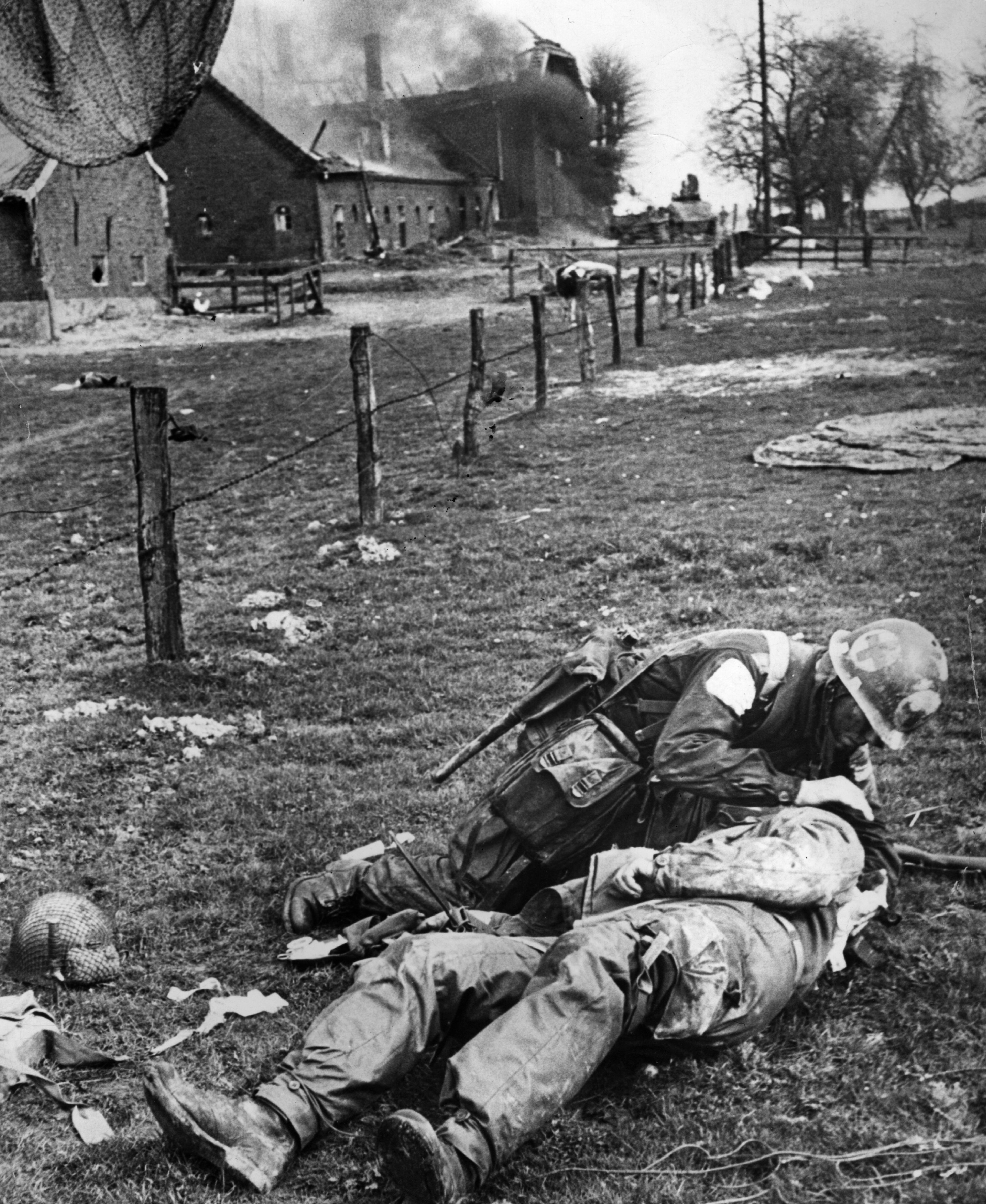 A medical orderly attending to an injured paratrooper on the ground, with a damaged building and fence in the background.