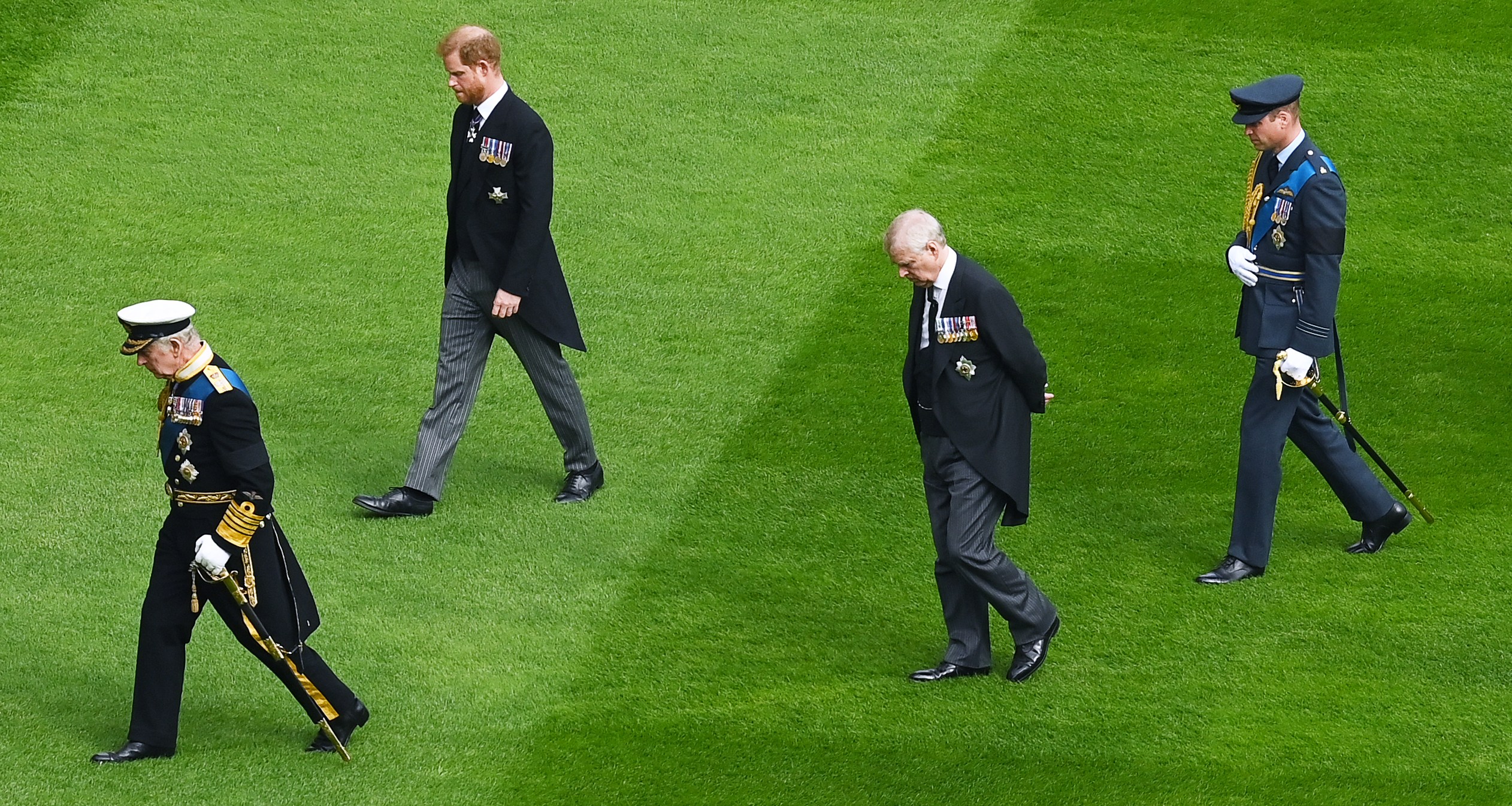 King Charles III, Prince Harry, Prince Andrew, and Prince William walk on green grass in military and ceremonial attire.