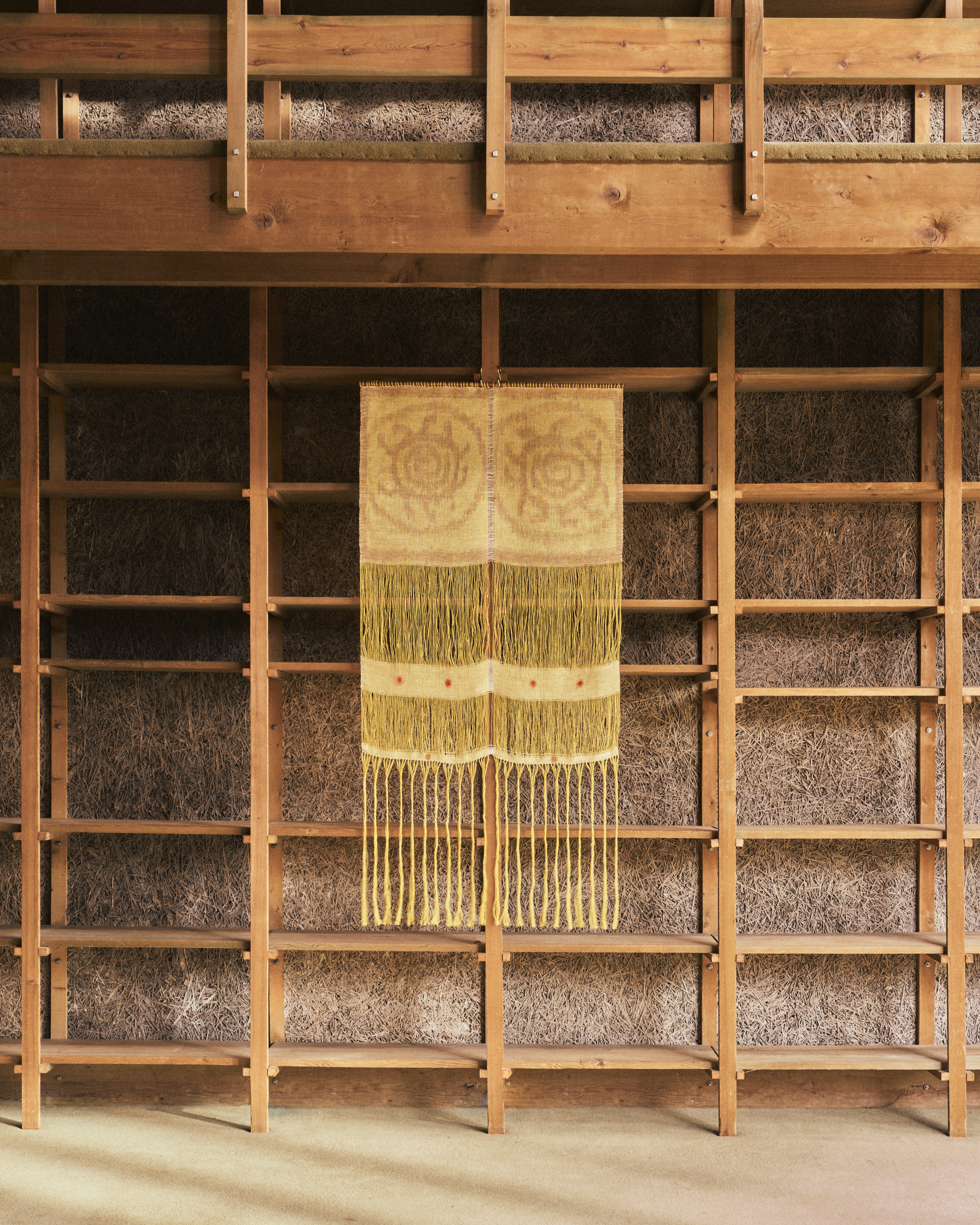 A yellow woven textile with rose patterns and fringe hanging in front of wooden shelves with hay-like back panels.