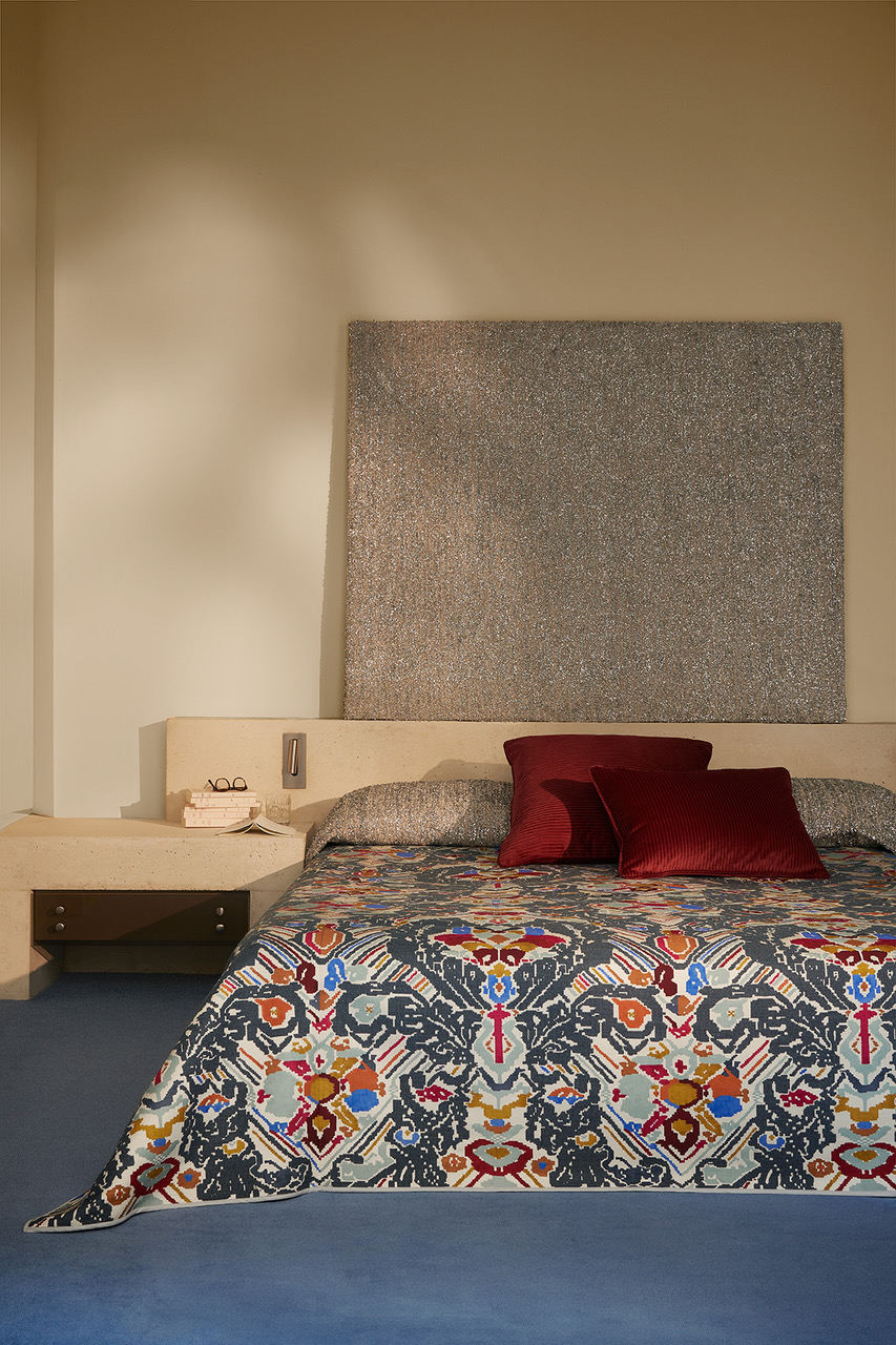 A bedroom featuring a bed with a colorful, patterned bedspread and red pillows, a beige headboard, and a textured silver art piece above it.