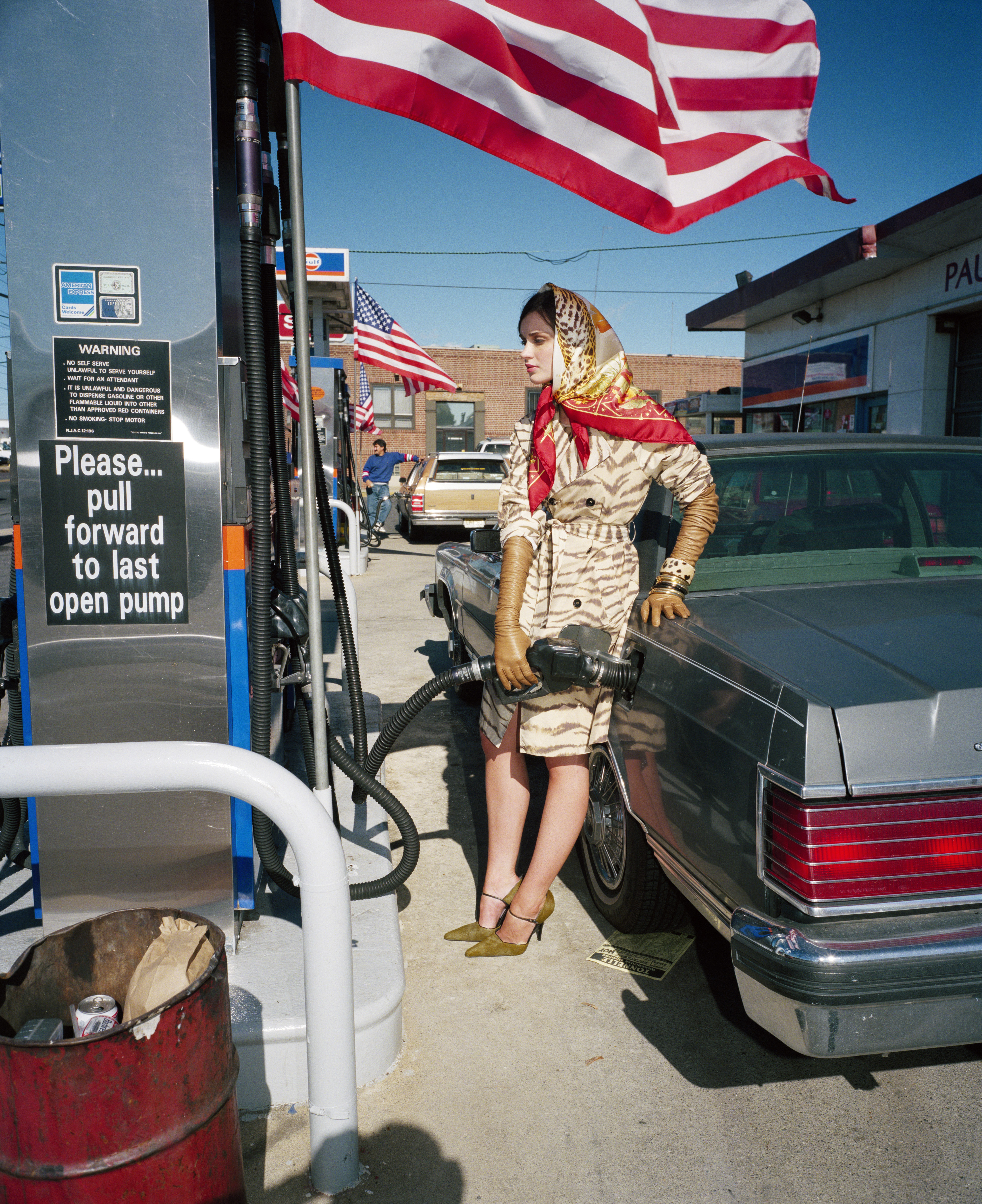 A fashion model in a tiger-print coat, gold gloves, and a red and gold headscarf pumps gas into a grey car at a gas station with an American flag flying above.