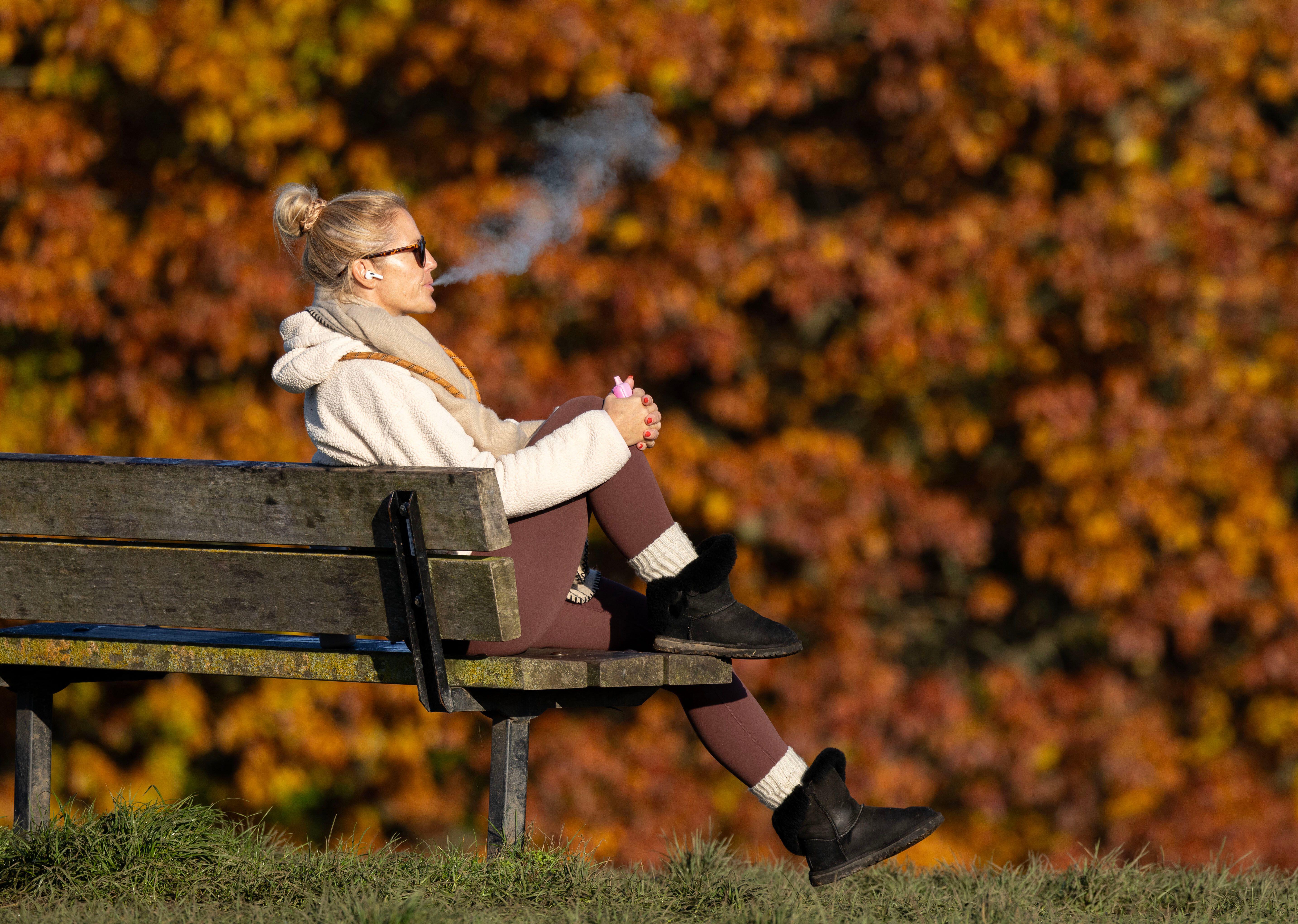 A woman exhales a plume of vapor while sitting on a park bench on Parliament Hill in London, with colorful autumnal trees in the background.