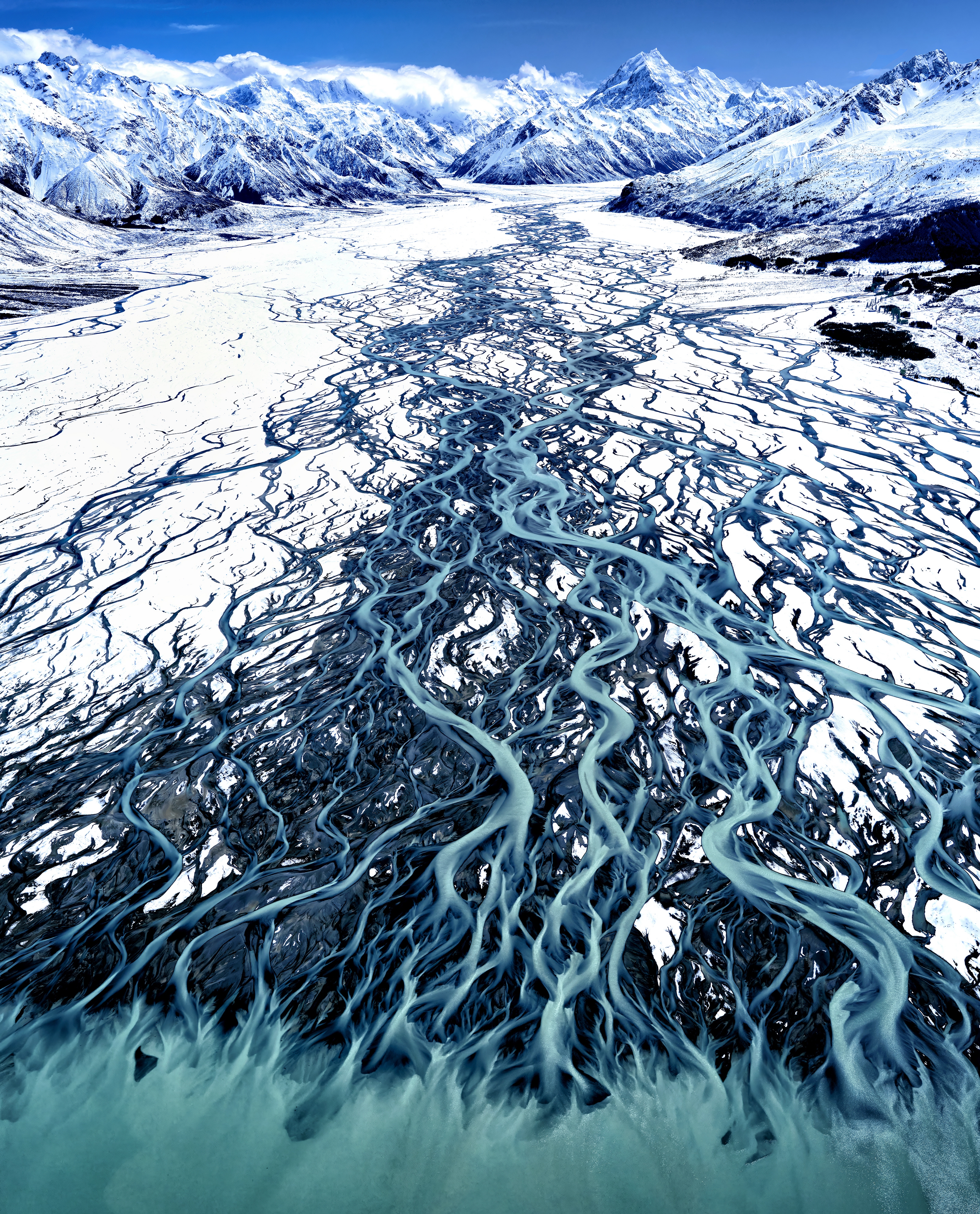 Aerial view of the Tasman River, a glacial river with many branching streams flowing into Lake Pukaki, surrounded by snowy mountains in New Zealand.