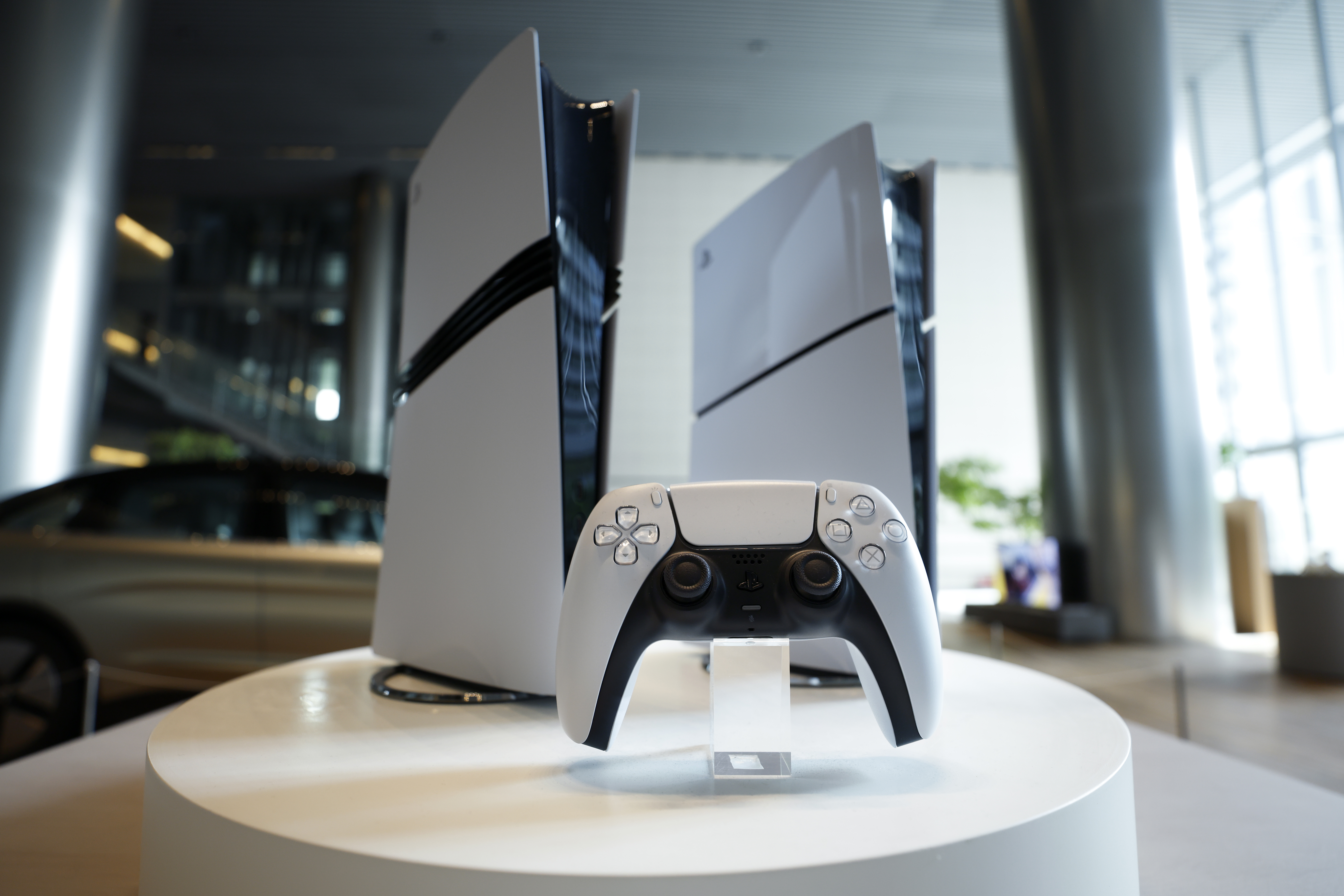 A white PlayStation 5 controller in front of a PS5 Pro and a PS5 Digital Edition console.