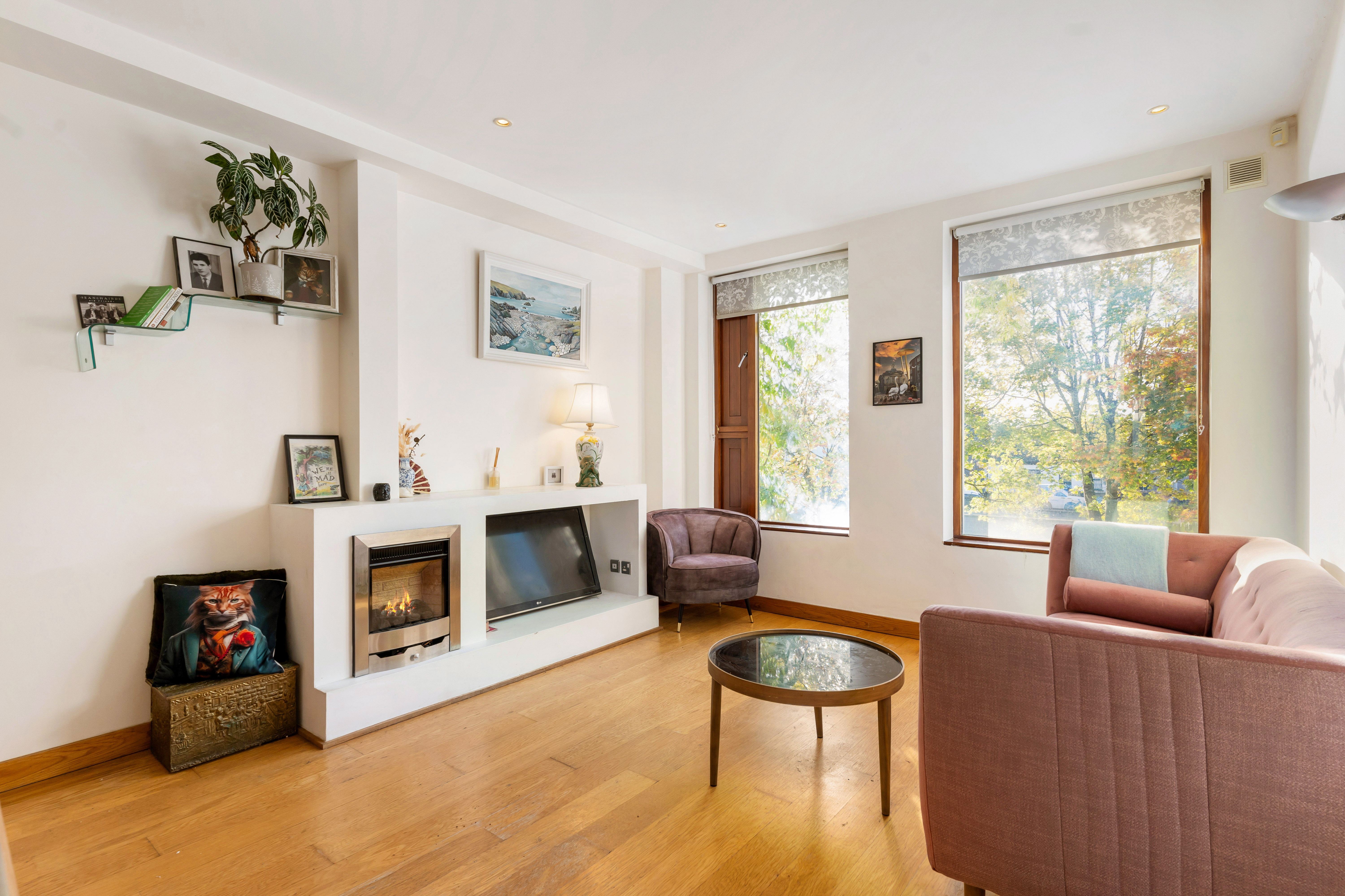 A well-lit living room with a fireplace, television, two couches, and large windows looking out onto trees.