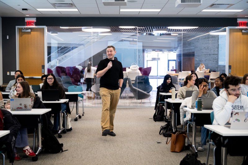 Matthias Schlichting, wearing a black sweater and tan pants, stands among his students, leading class. Behind him is a glass wall looking into Northeastern's ISEC building.