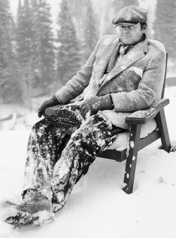 A man in a suit, gloves, and flat cap sits on a wooden chair outdoors, covered in snow, with snow-covered trees in the background. He appears cold and his clothes are dusted with snow.
