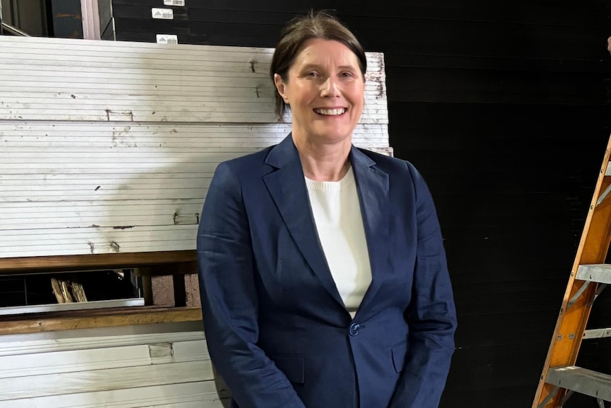 Robyn, wearing a navy suit with white shirt, smiles as she leans against a stack of solar panels